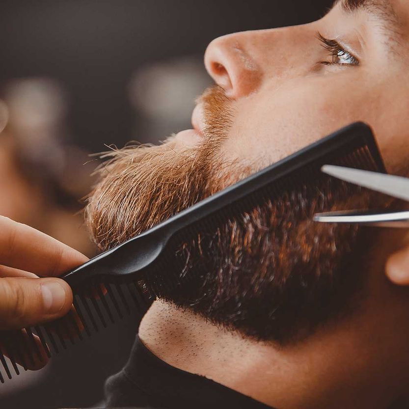Hipster-Mann sitzt im Sessel-Friseursalon, während der Friseur den Bart mit einer Schere rasiert.